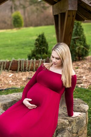 Pregnant woman in red off-shoulder gown cradling baby bump outdoors near stone wall and green garden.
