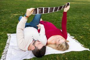 Expecting couple lying on blanket in park holding ultrasound photos, celebrating pregnancy together outdoors.