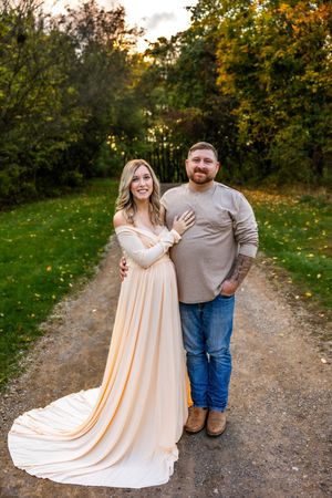 Couple posing on a dirt path surrounded by autumn trees, woman in elegant peach gown, man in casual jeans and tan shirt.