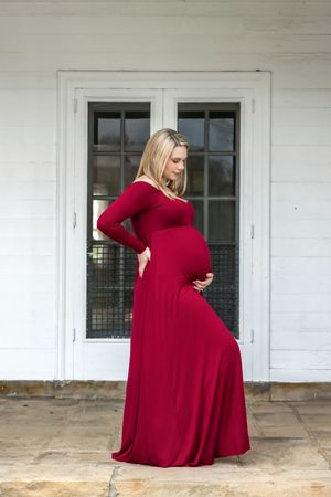 Pregnant woman in elegant red maxi dress posing outdoors against white building with French doors.
