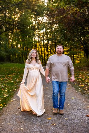 Pregnant couple holding hands walking on a tree-lined path in autumn, woman in elegant cream gown, man in casual jeans.
