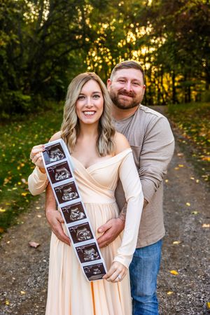 Smiling couple holding ultrasound photos on a forest path during golden hour, announcing their pregnancy.