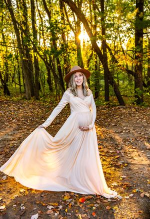 Pregnant woman in flowing cream gown and brown hat poses in autumn forest with golden sunlight streaming through trees.