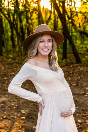 Pregnant woman in cream off-shoulder dress and brown hat cradling baby bump in autumn forest.