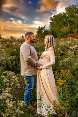 Couple shares tender moment in wildflower field at sunset, woman in elegant cream maternity gown cradling baby bump.