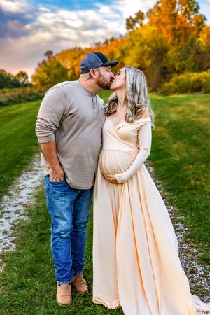 Couple sharing a kiss outdoors during golden hour, pregnant woman in flowing cream gown cradling baby bump, man in jeans and cap.