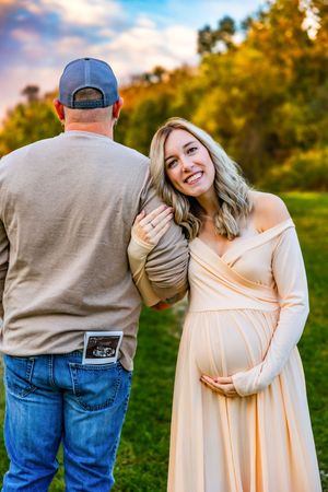 Smiling pregnant woman in cream dress holds baby bump beside man with sonogram photo in jeans pocket outdoors.