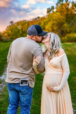 A couple shares a kiss outdoors during a maternity photo shoot, with an ultrasound photo tucked in his back pocket.