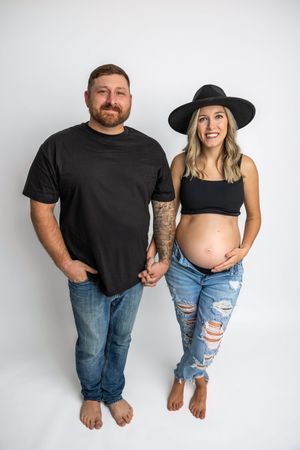 Couple posing for maternity photo, man in black tee and jeans, pregnant woman in crop top, ripped jeans, and black hat.