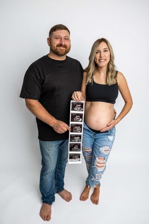 Expecting couple holding ultrasound photos, woman in crop top and ripped jeans showing baby bump, man in black tee, white background.