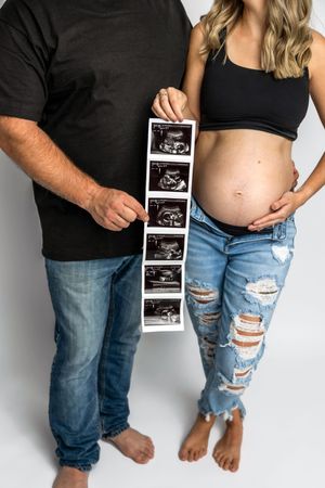 Pregnant couple holding ultrasound sonogram strip, woman in ripped jeans cradling baby bump, maternity announcement photo.