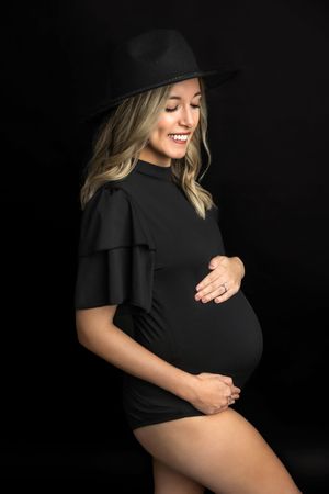 Smiling pregnant woman in black ruffle dress and wide-brim hat cradling baby bump against dark background.