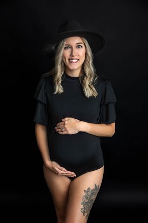 Smiling pregnant woman in black bodysuit and wide-brim hat with floral thigh tattoo poses against dark background.