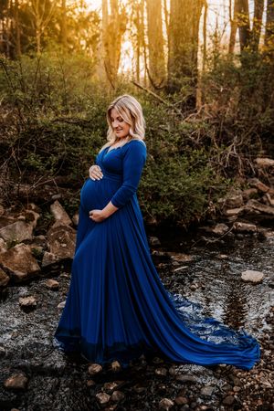 Pregnant woman in royal blue maternity gown standing by a rocky stream in golden hour forest light.