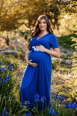 Pregnant woman in blue maxi dress holding baby bump, surrounded by blue iris flowers in a sunlit garden.