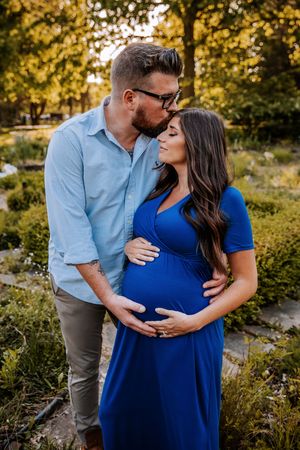 A man kisses a pregnant woman in a blue dress outdoors, both cradling her baby bump in a lush garden setting.