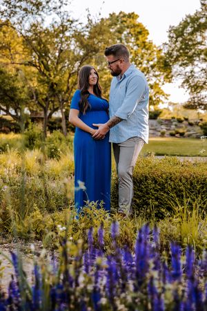 Pregnant couple holding hands in a garden with purple flowers, woman in blue maternity dress, golden hour lighting.