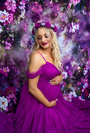 Pregnant woman in purple tulle gown and floral crown poses gracefully against a vibrant floral backdrop.