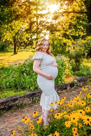 Pregnant woman in floral dress cradling baby bump among yellow black-eyed Susans at golden hour in lush garden.