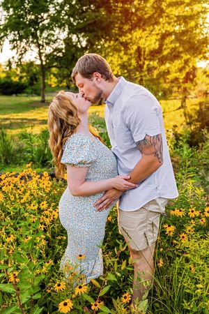 Pregnant couple kissing in a sunny garden surrounded by yellow black-eyed Susan flowers during golden hour.