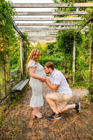 A man kneels to kiss a pregnant woman's belly under a wooden pergola surrounded by lush greenery on a brick path.