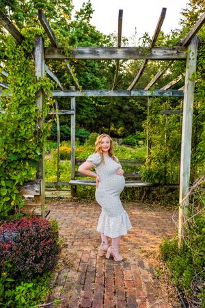 Pregnant woman in white dress posing under wooden pergola surrounded by lush green garden foliage on brick pathway.