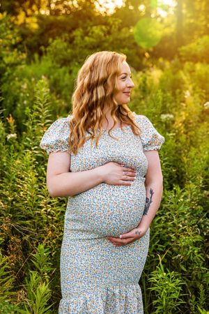 Pregnant woman in floral dress cradling baby bump outdoors in golden sunlight surrounded by lush green foliage.