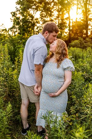 A couple shares a tender kiss at golden hour in a lush green field, with the woman in a floral dress cradling her baby bump.