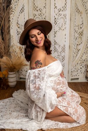 Pregnant woman in white lace off-shoulder dress and brown hat, sitting gracefully in a boho-styled maternity photo shoot.