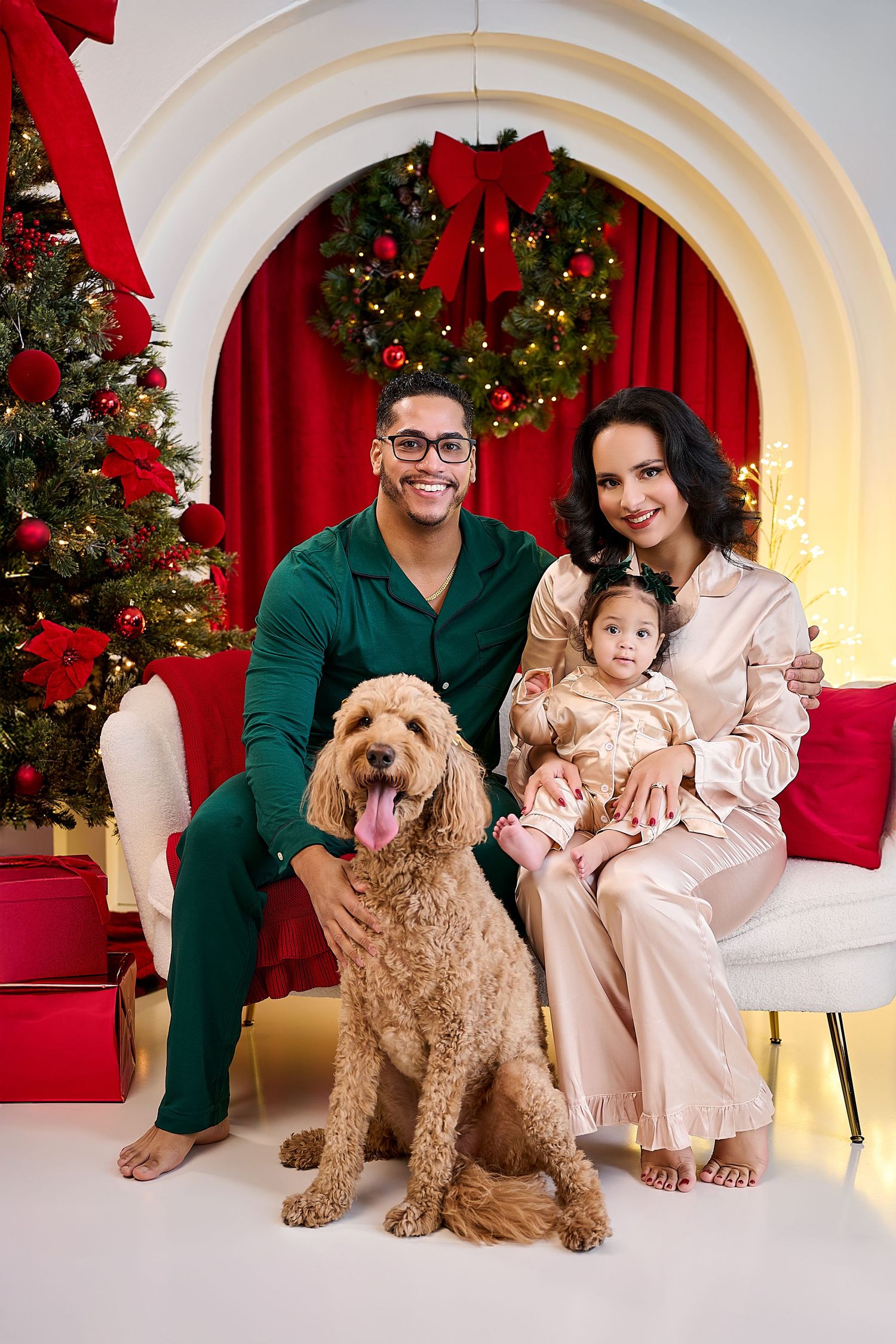 Family in festive pajamas poses with golden doodle dog in front of Christmas tree and holiday wreath.