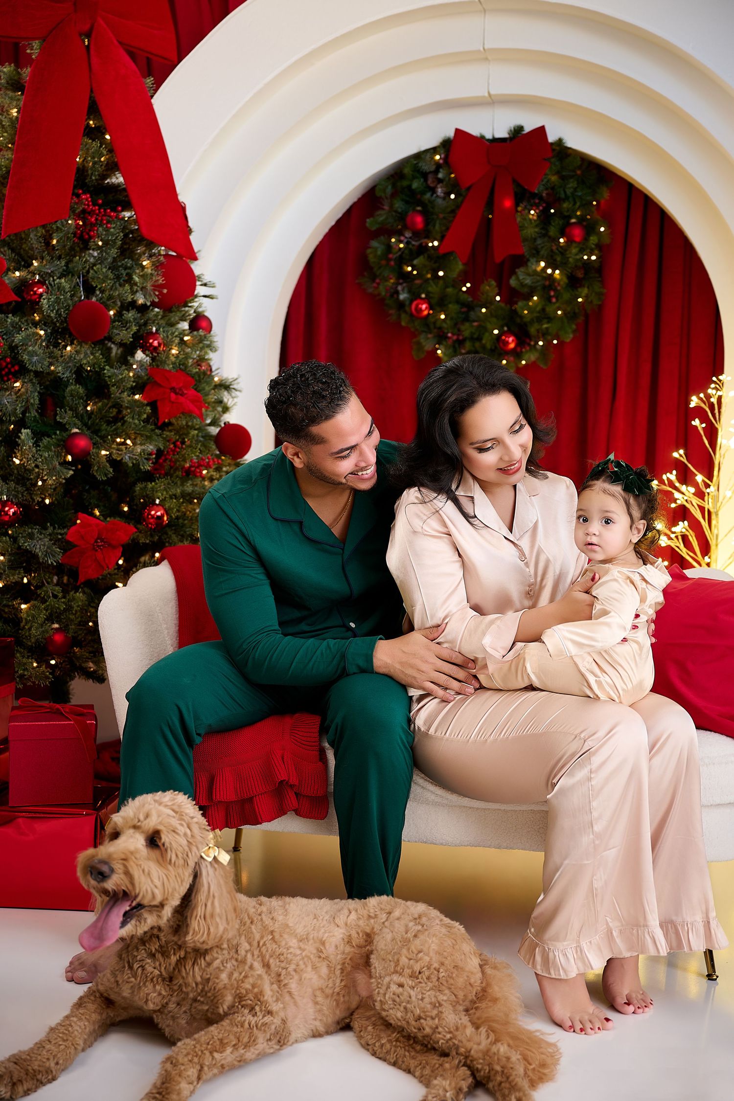 Family in satin pajamas poses with golden doodle dog beside decorated Christmas tree and festive wreath backdrop.