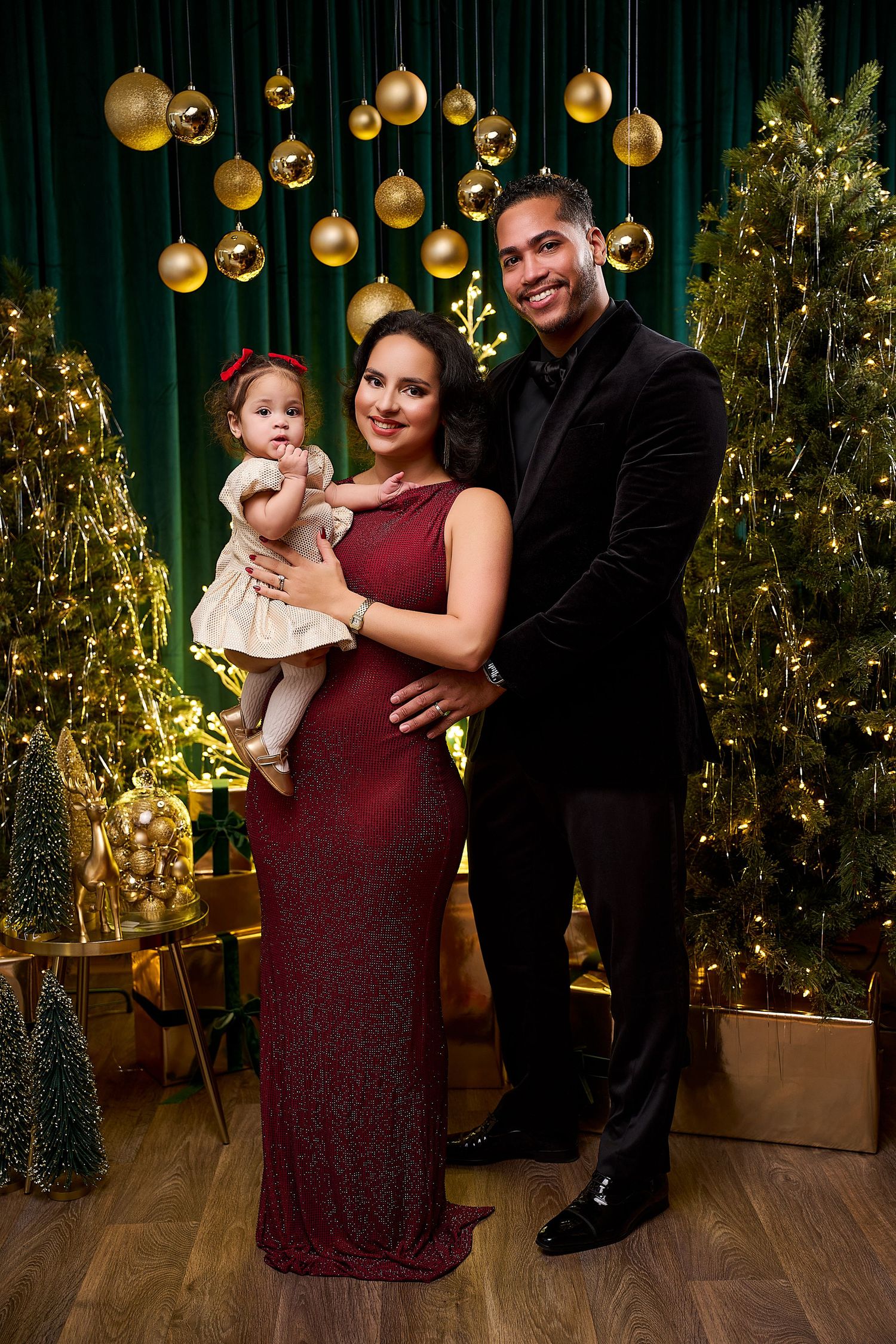 Elegant family Christmas portrait with mother in red gown, father in black suit, holding baby girl amid golden ornaments.