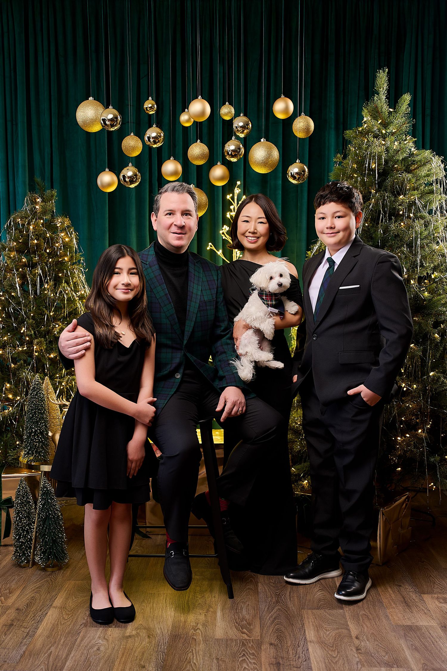 Family of four with a small white dog posing in front of Christmas trees and gold ornaments on green backdrop.