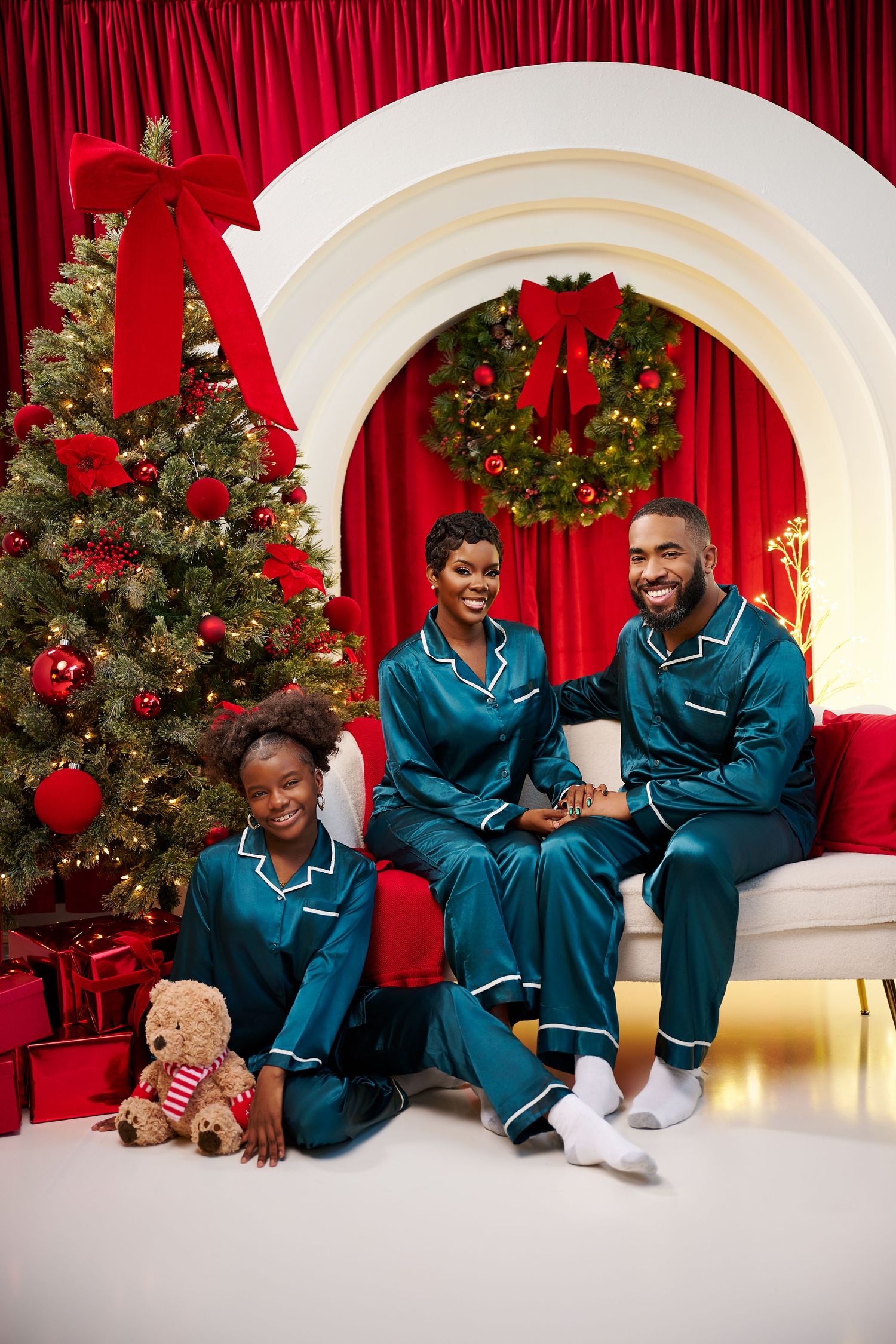 Family of three in matching teal satin pajamas posing by a decorated Christmas tree with red gifts and holiday wreath.