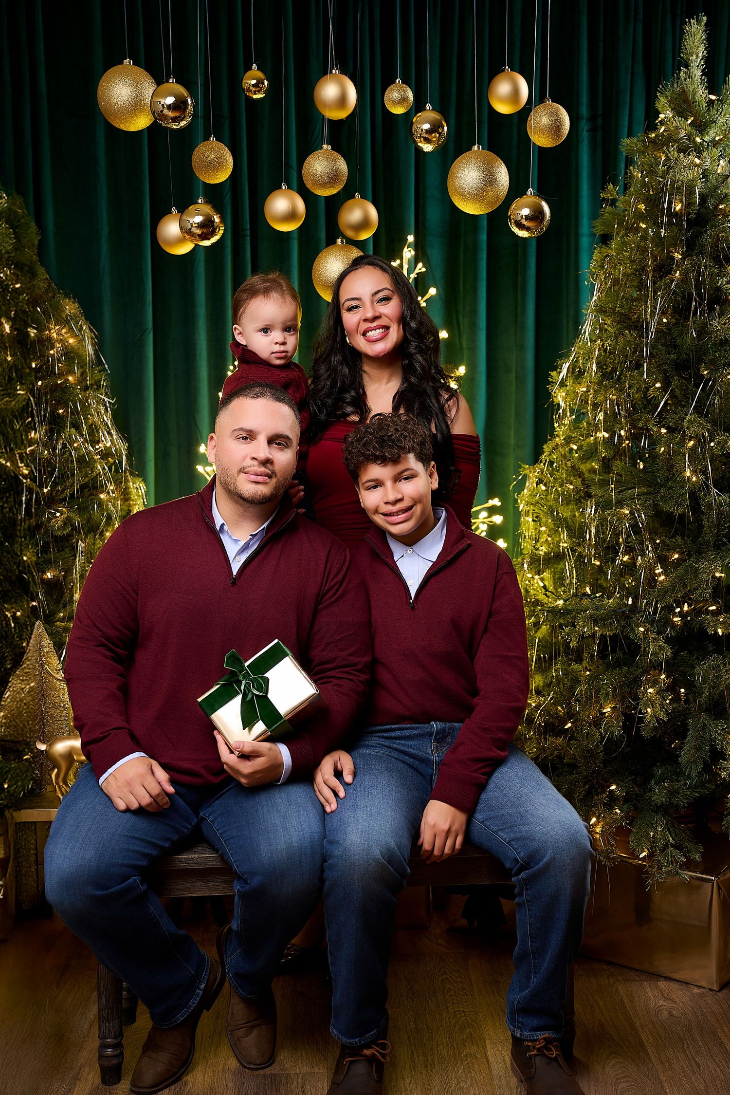 Family of four in matching maroon outfits poses with Christmas trees and gold ornaments in a festive holiday portrait.