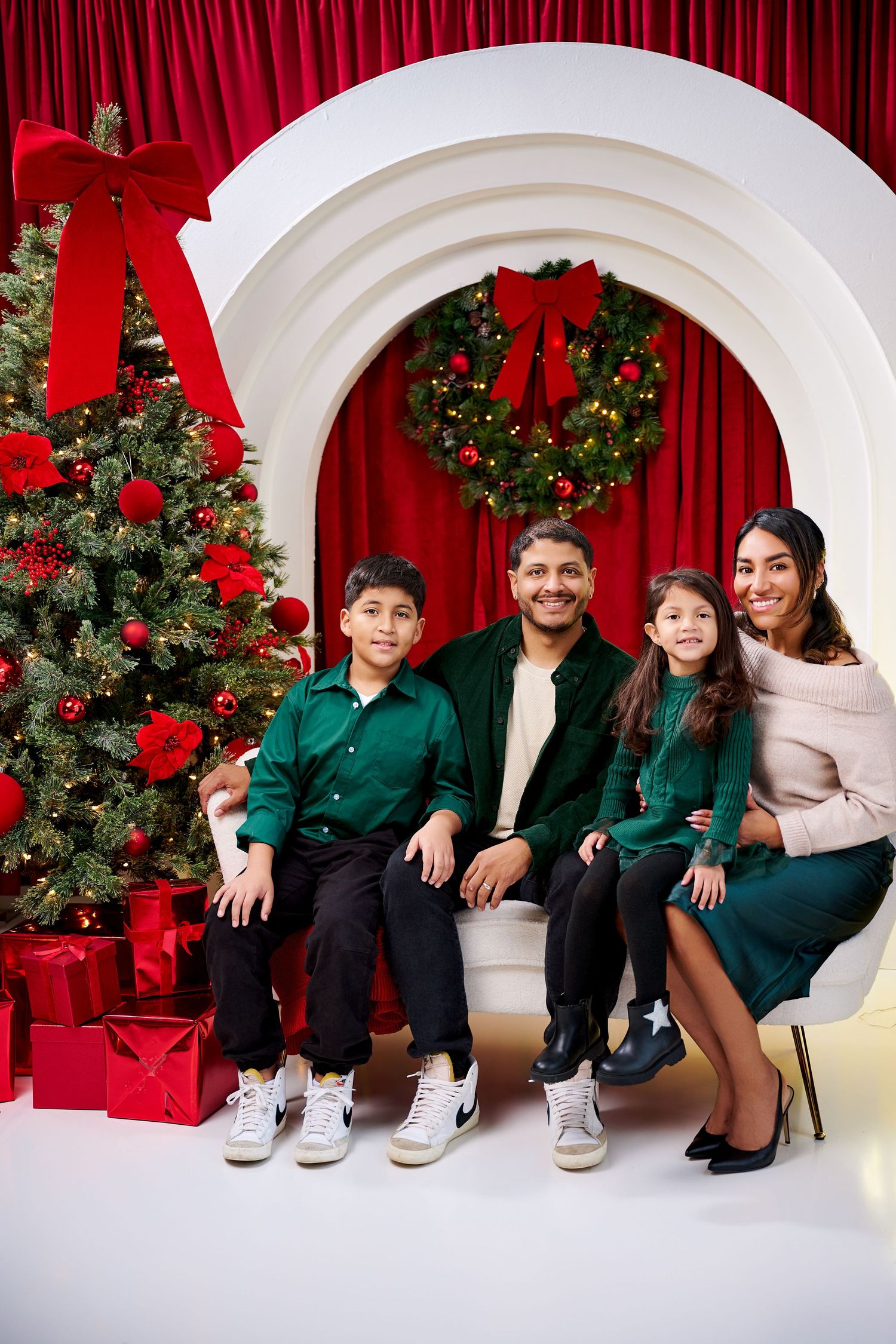 Family of four posing for a Christmas portrait with decorated tree, wreath, and red curtain backdrop.