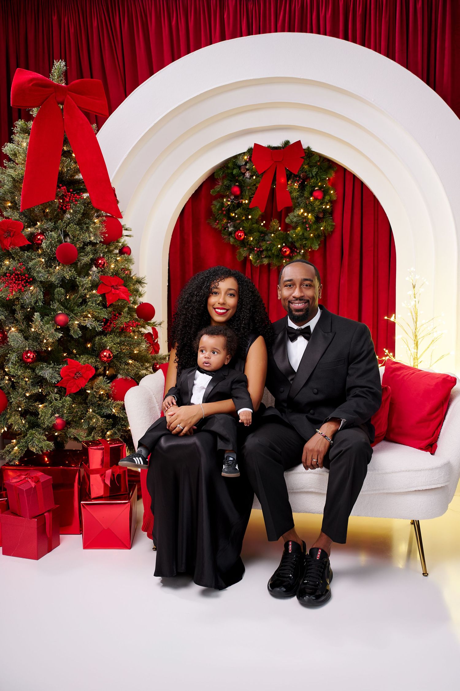 A family of three in elegant black attire poses for a Christmas portrait with a decorated tree and red holiday backdrop.