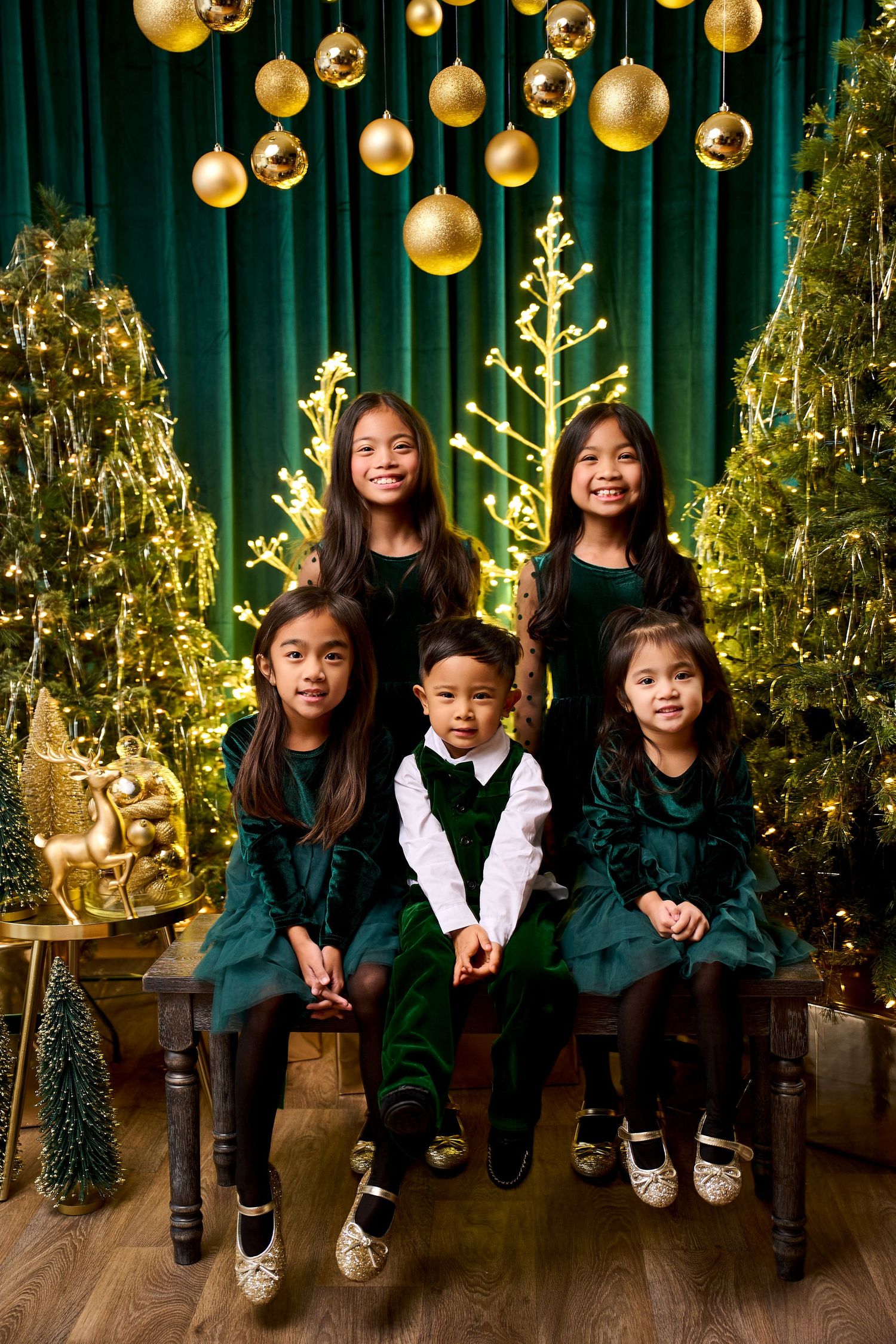 Five children in green velvet holiday outfits pose together among Christmas trees and gold ornaments.