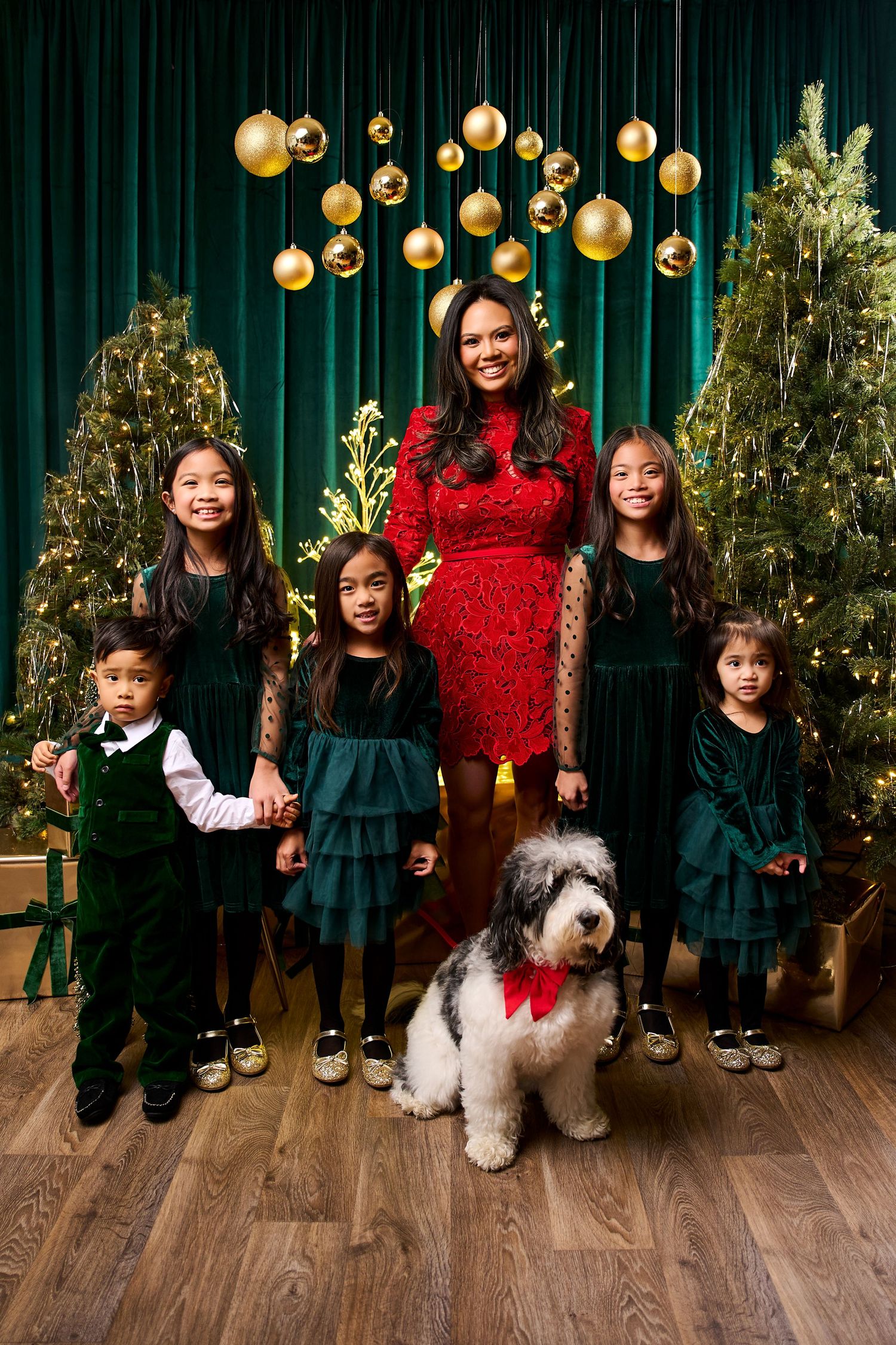Family Christmas photo with woman in red dress, four children in green outfits, and a fluffy dog with red bow by decorated trees.