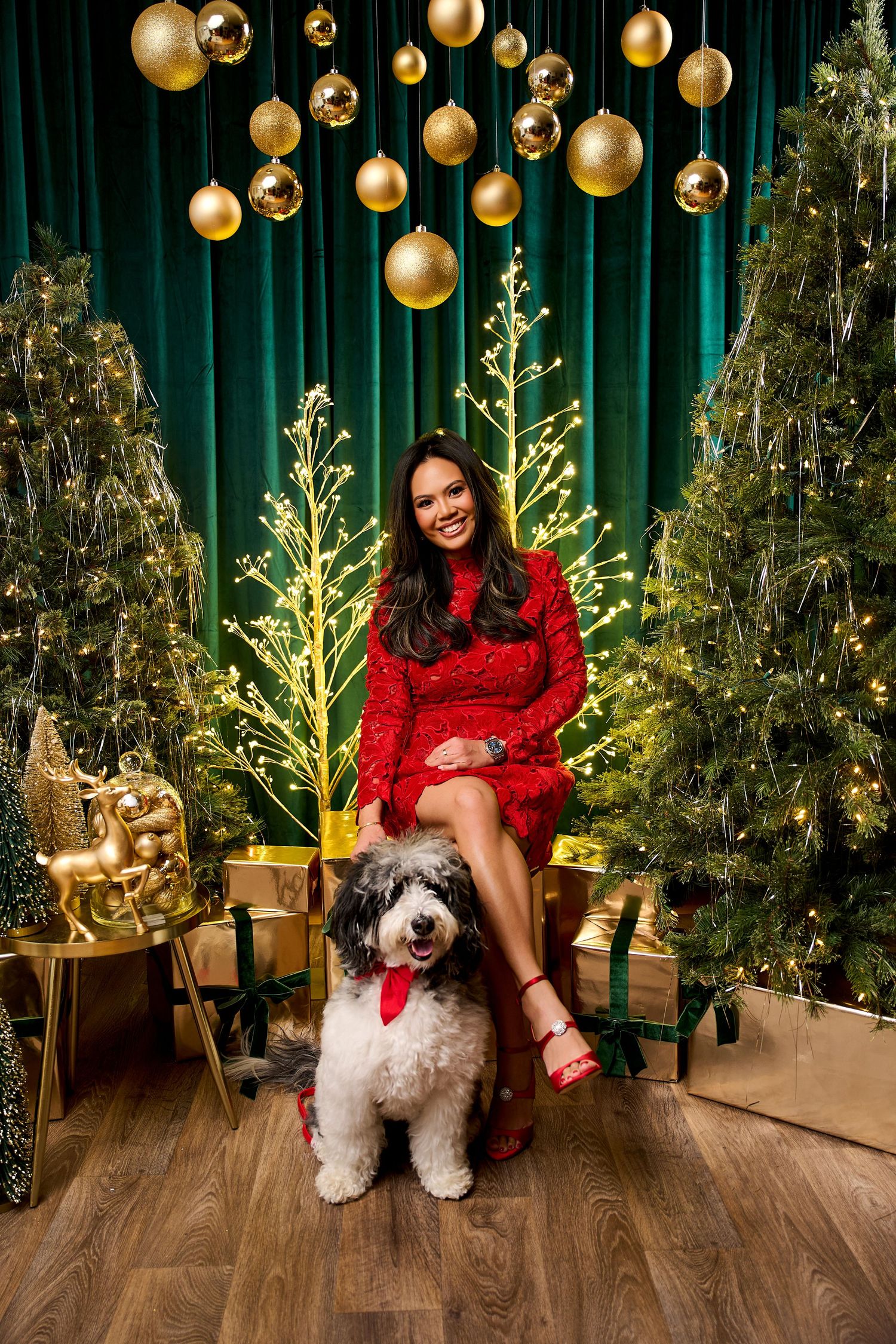 Woman in red dress smiling with fluffy dog wearing tie, surrounded by Christmas trees and gold ornaments.