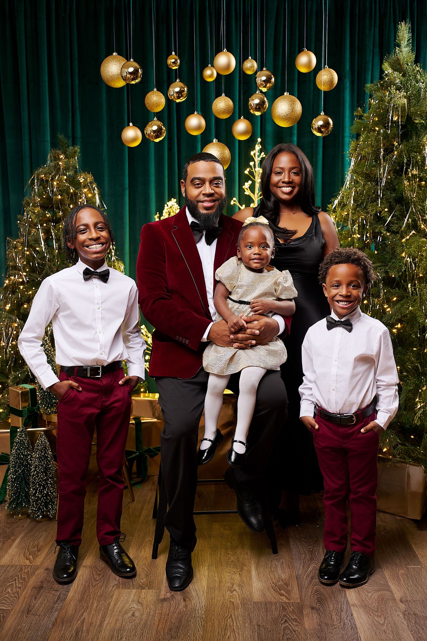 A joyful family of five poses in elegant holiday attire before decorated Christmas trees with gold ornaments.