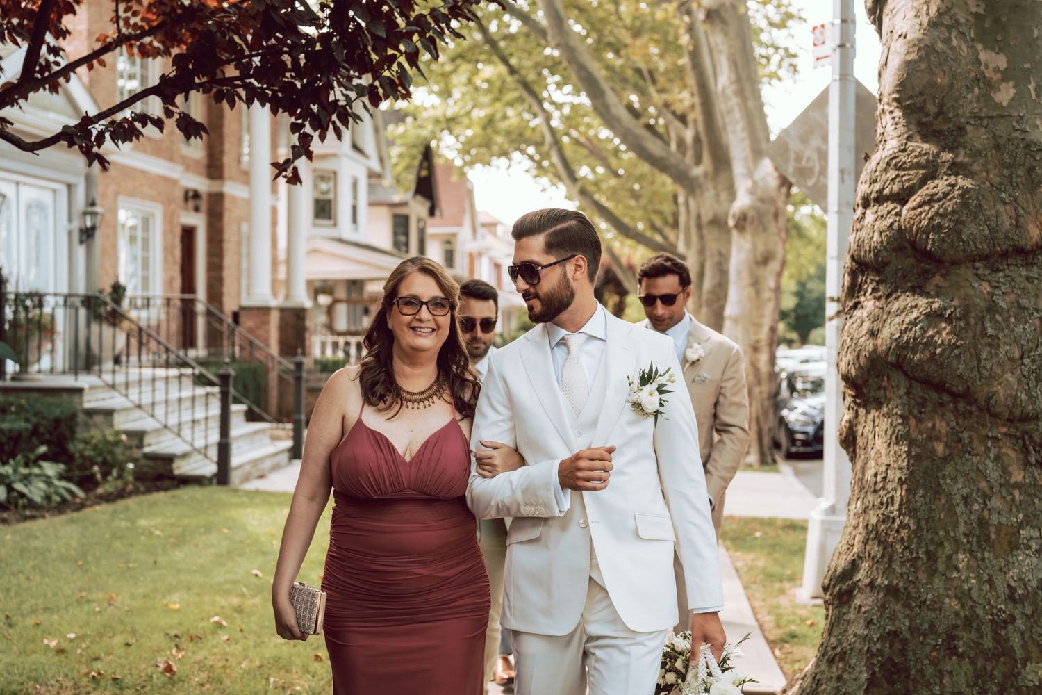 A groom in a white suit walks with a bridesmaid in a burgundy dress on a tree-lined suburban sidewalk.
