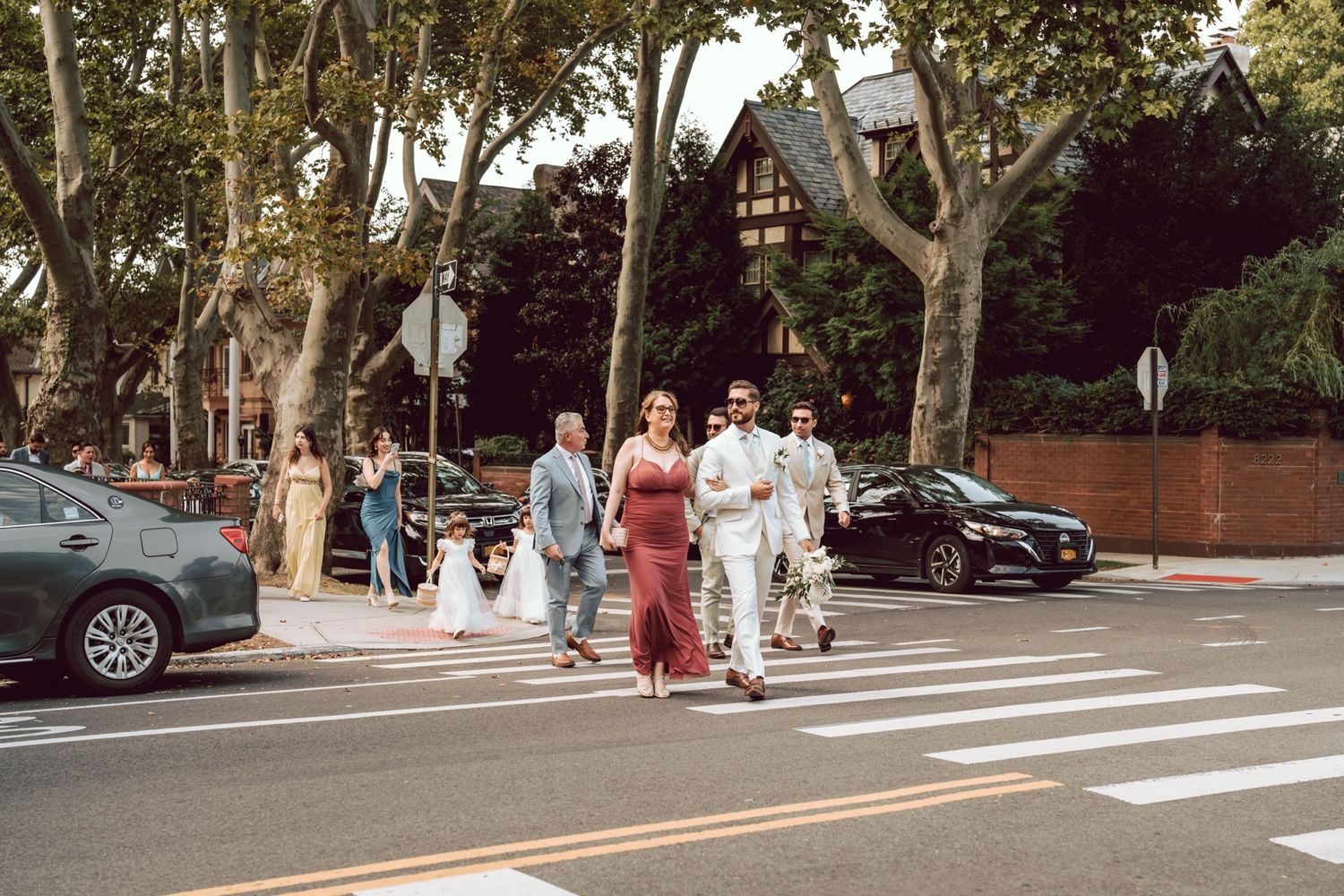 Wedding party crosses a tree-lined urban street, bride in white gown with guests in formal attire on a sunny day.