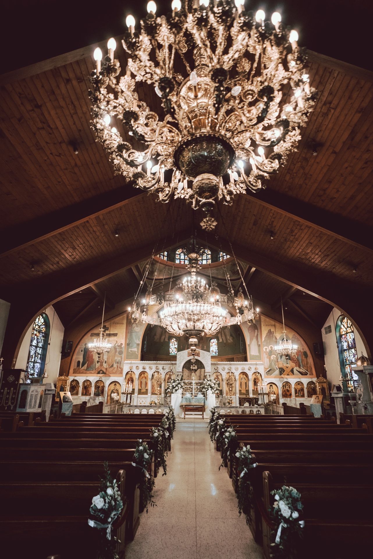 Ornate Orthodox church interior with grand chandelier, wooden pews, and floral decorations along the aisle for a wedding ceremony.