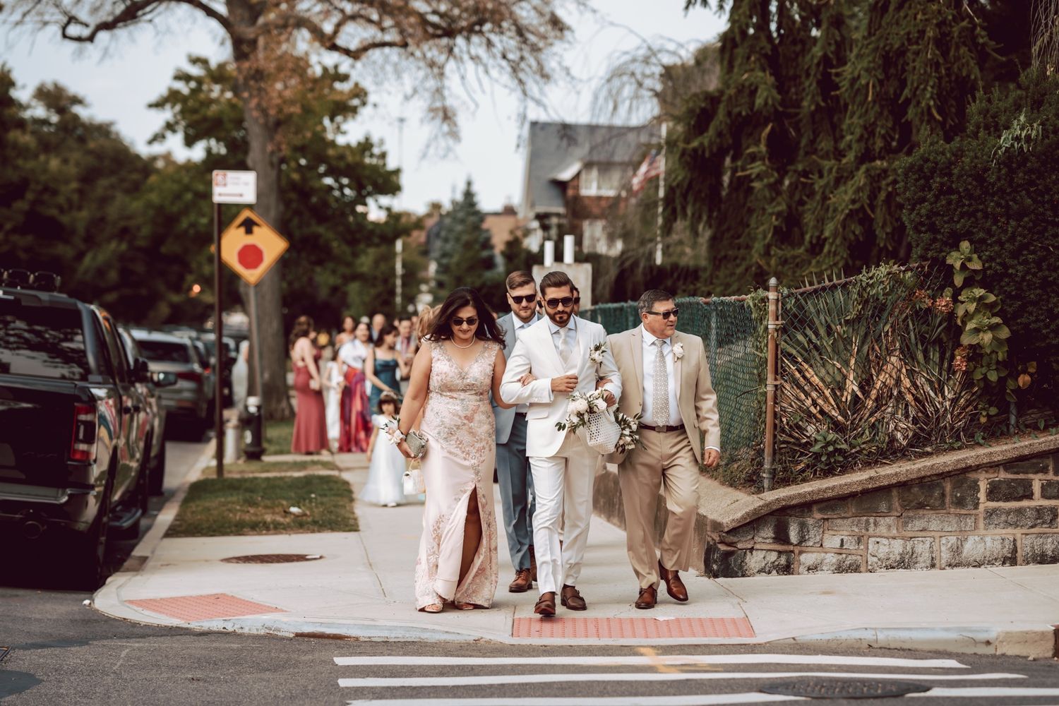 Wedding party walks along a sunny suburban sidewalk, bride in pink gown, groom in white suit holding bouquet.