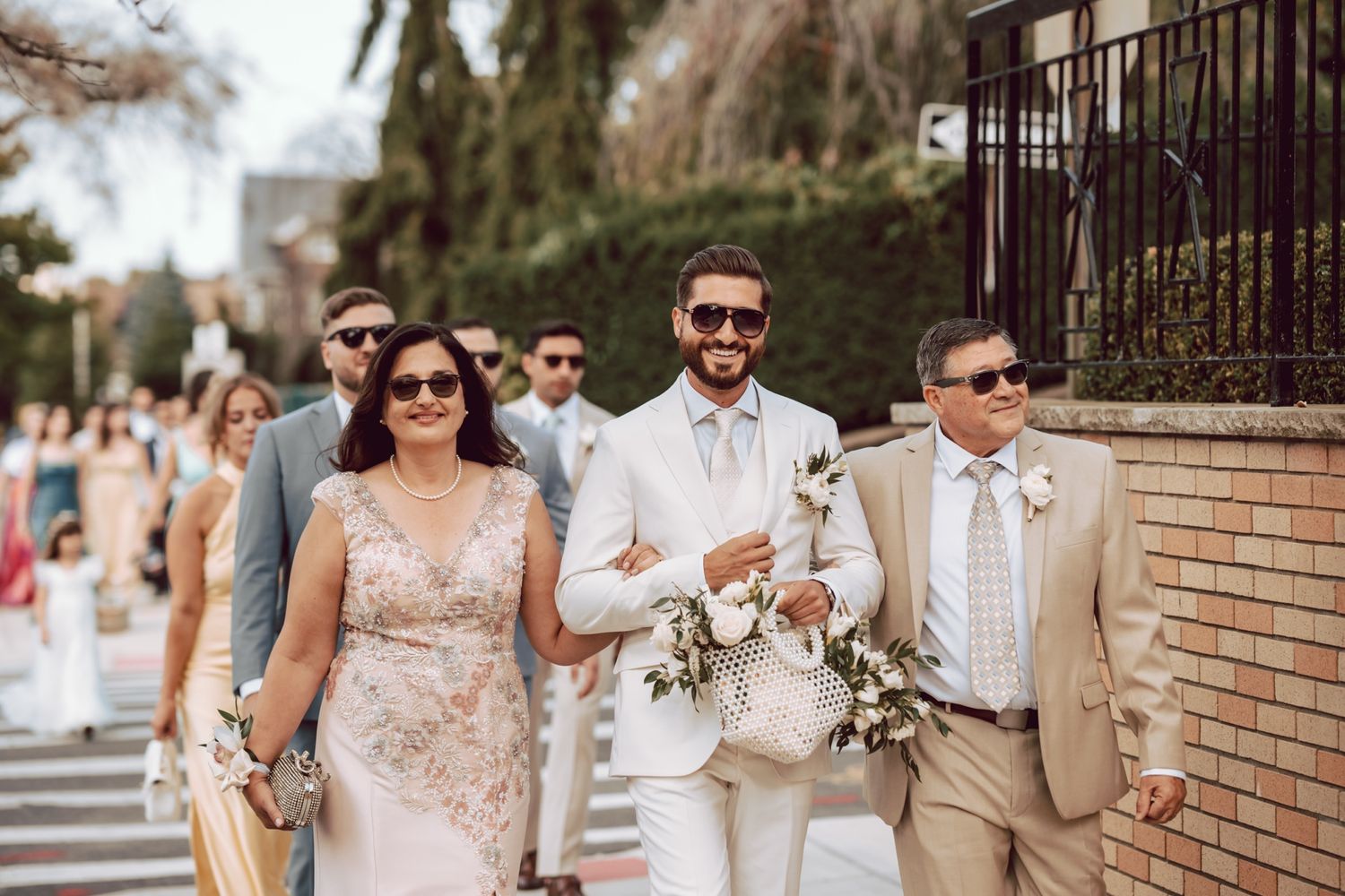 Groom in white suit walks with family during outdoor wedding procession, flanked by elegantly dressed guests.