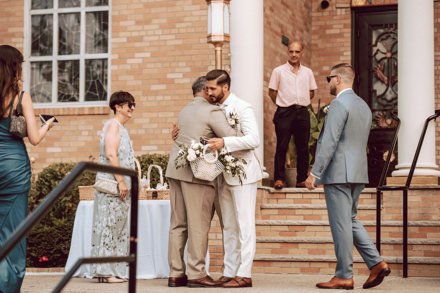 Two grooms share a heartfelt hug outside a brick wedding venue surrounded by elegantly dressed guests.