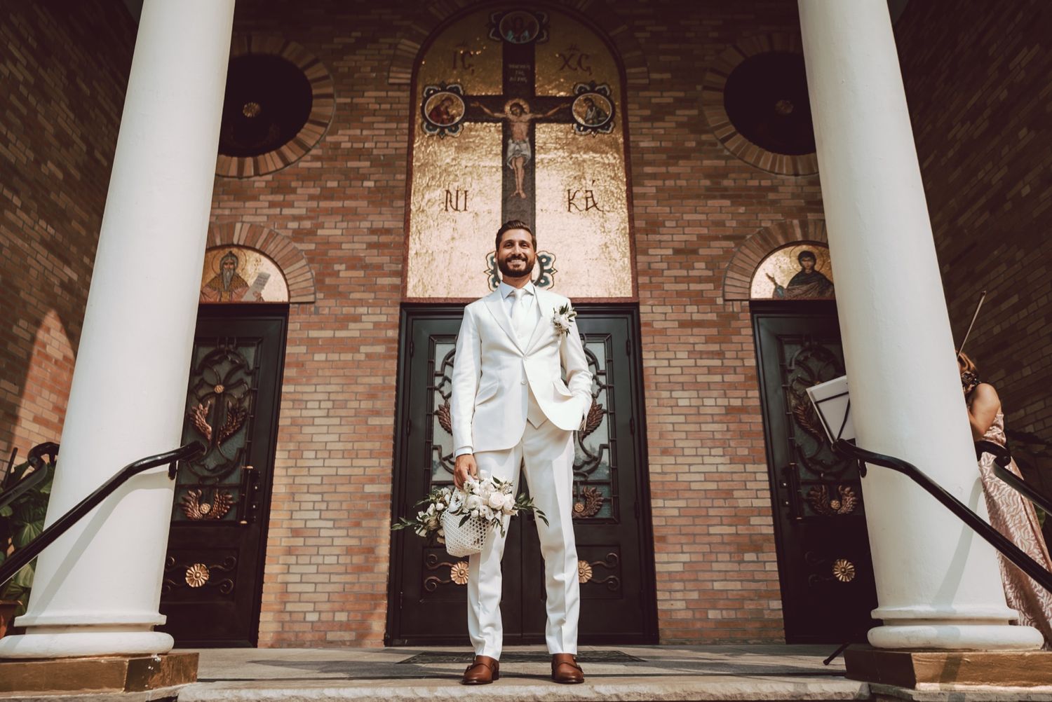 Groom in white suit holding bouquet stands on church steps with ornate cross and brick facade backdrop.