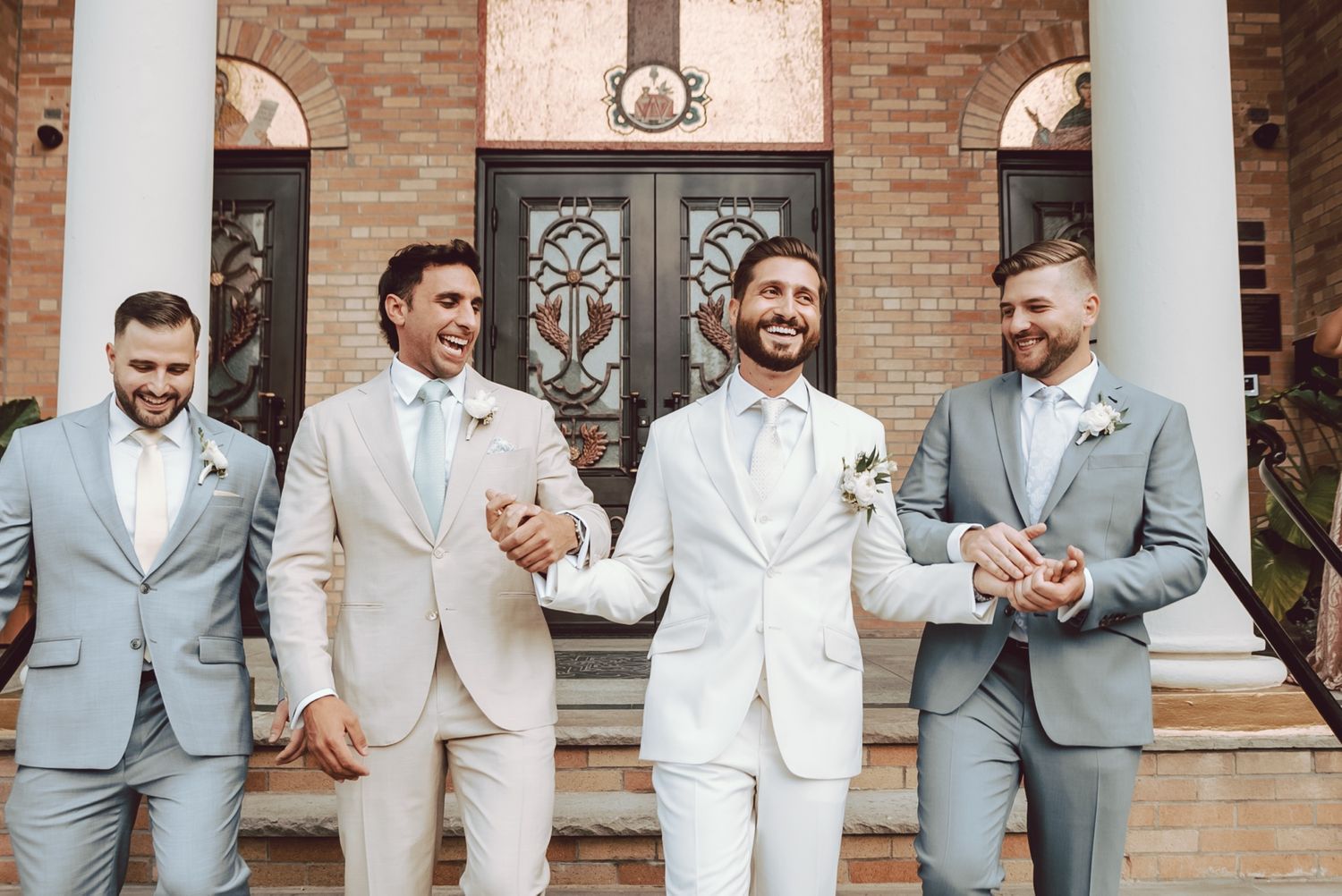 Groom in white suit walks with three groomsmen in gray suits outside elegant brick building.