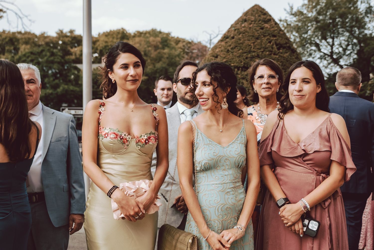 Three elegantly dressed women standing together outdoors at a wedding reception, wearing formal gowns in gold, blue, and pink.